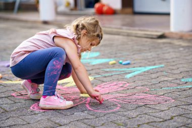 Bahçede renkli tebeşir çiçekleriyle resim yapan küçük bir anaokulu kızı. Mutlu çocuk asfaltta resim çiziyor ve resimler yaratıyor. Yazın yaratıcı açık hava çocuk aktivitesi.
