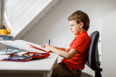 Gözlüklü küçük bir çocuğun portresi. Ev ödevi yapıyor, yazıyor ve öğreniyor. Küçük çocuk içeride egzersiz yapıyor. İlkokul ve eğitim, evde eğitim kavramı..