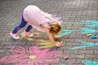 Bahçede renkli tebeşir çiçekleriyle resim yapan küçük bir anaokulu kızı. Mutlu çocuk asfaltta resim çiziyor ve resimler yaratıyor. Yazın yaratıcı açık hava çocuk aktivitesi.