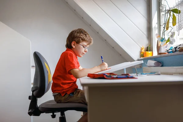 Porträtt av en liten pojke med glasögon hemma som gör läxor, skriver och lär. Ett litet barn som motionerar inomhus. Grundskola och utbildning, hemskolekoncept. — Stockfoto