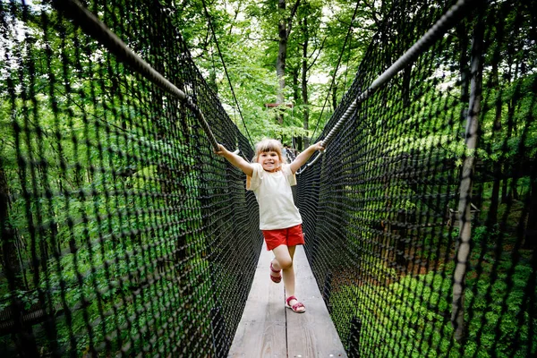 Klein kleuter meisje wandelen op hoge boom-bladerdak pad met houten loopbrug en touwen op Hoherodskopf in Duitsland. Gelukkige actieve kind verkennen boomtop pad. Grappige activiteit voor gezinnen buiten — Stockfoto