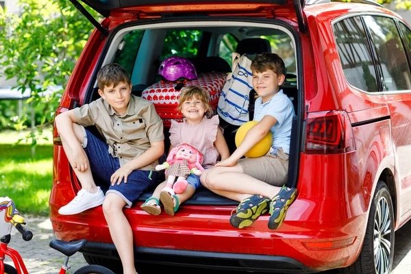 Tři děti, dva chlapci a školačka sedící v kufru auta před odjezdem na letní prázdniny s rodiči. Šťastné děti, sourozenci, bratři a sestry s kufry a hračkami na cestu — Stock fotografie