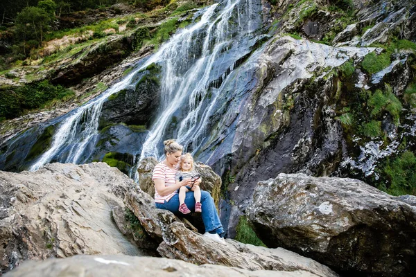 Linda niña y madre sentadas cerca de la cascada de agua de Powerscourt Waterfall, la cascada más alta de Irlanda en co. Wicklow. Vacaciones familiares con niños pequeños. Mujer y bebé niño — Foto de Stock