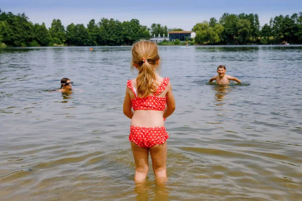 เด็กสาวผมบลอนด์เล็ก ๆ น้อย ๆ มีความสนุกสนานกับการกระเด็นในทะเลสาบในวันฤดูร้อนกลางแจ้ง เด็กที่มีความสุขเรียนรู้การว่ายน้ํา พักผ่อนหย่อนใจกับเด็กในวันหยุด อันตรายที่ทะเลสาบภายในประเทศ — ภาพถ่ายสต็อก