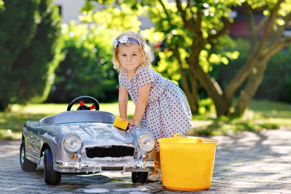 Menina bonito lindo bebê lavando grande carro de brinquedo velho no jardim de verão, ao ar livre. Feliz saudável pequena criança limpando carro com água e sabão, se divertindo com espirrar e brincar com esponja. — Fotografia de Stock