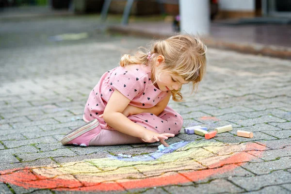 Pequeña niña preescolar pintando arco iris con tiza de colores en el suelo en el patio trasero. Positivo feliz niño pequeño dibujo y la creación de imágenes. Actividad creativa al aire libre en verano. — Foto de Stock