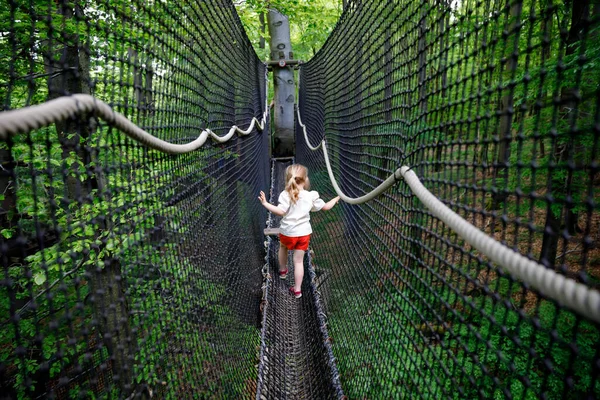 Cute little preschool girl walking on high tree-canopy trail with wooden walkway and ropeways on Hoherodskopf in Germany. Happy active child exploring treetop path. Fun activity for families outdoors — Fotografia de Stock