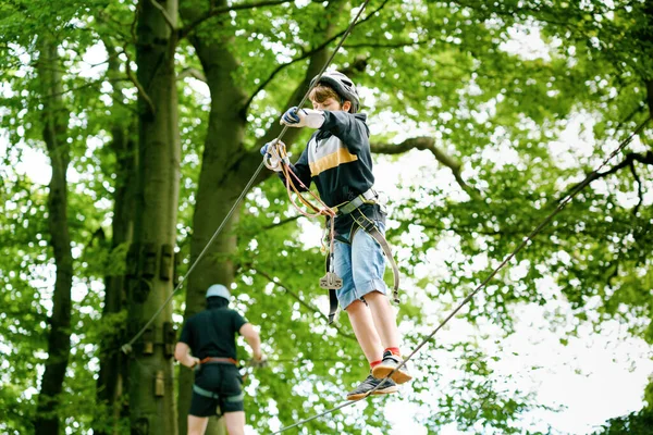 School boy in forest adventure park. Acitve child, kid in helmet climbs on high rope trail. Agility skills and climbing outdoor amusement center for children. Outdoors activity for kid and families.