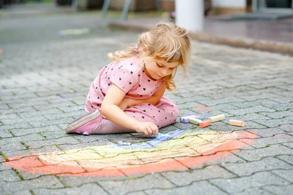 裏庭にカラフルなチョークで虹を描く小さな就学前の女の子。肯定的な幸せな幼児の子供の絵を描き、絵を作成する。夏の創造的な屋外活動. — ストック写真