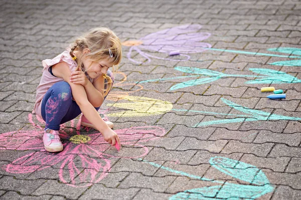 Bahçede renkli tebeşir çiçekleriyle resim yapan küçük bir anaokulu kızı. Mutlu çocuk asfaltta resim çiziyor ve resimler yaratıyor. Yazın yaratıcı açık hava çocuk aktivitesi. — Stok fotoğraf