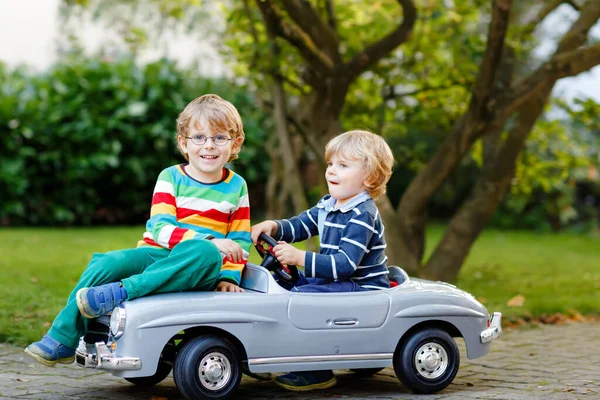Δύο μικρά παιδιά προσχολικής ηλικίας παίζουν με ένα μεγάλο παλιό αυτοκινητάκι στον καλοκαιρινό κήπο, σε εξωτερικούς χώρους. Ευτυχισμένα παιδιά παίζουν μαζί, οδηγώντας αυτοκίνητο. Εξωτερική δραστηριότητα για παιδιά. Αδέρφια και φίλοι τη ζεστή μέρα — Φωτογραφία Αρχείου
