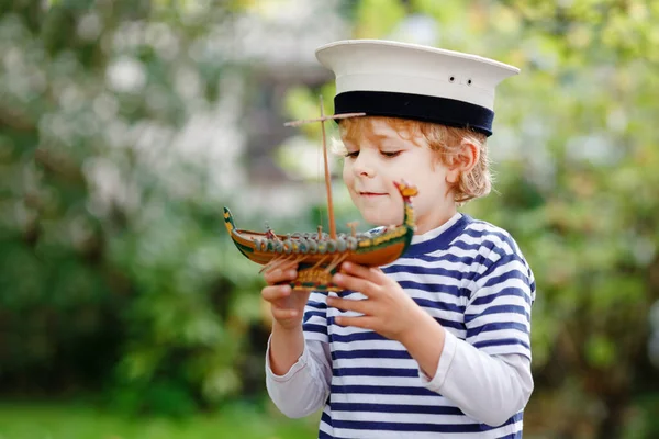 Boldog kisfiú tengerész sapkában és egyenruhában, aki matrózhajóval játszik. Mosolygó óvodás gyerek álmodik és szórakozik. Oktatás, szakma, álomkoncepció — Stock Fotó