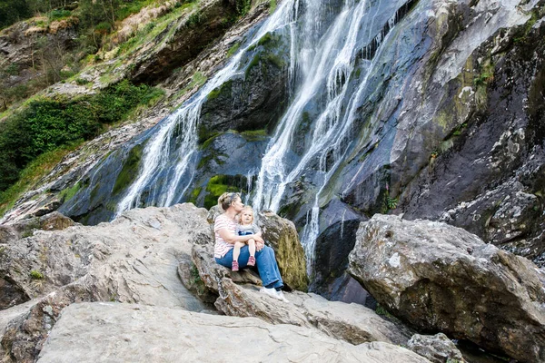 Linda niña y madre sentadas cerca de la cascada de agua de Powerscourt Waterfall, la cascada más alta de Irlanda en co. Wicklow. Vacaciones familiares con niños pequeños. Mujer y bebé niño — Foto de Stock