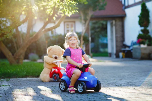 Pequena menina adorável criança dirigindo carro de brinquedo e se divertindo com brincar com urso de brinquedo de pelúcia, ao ar livre. Linda criança saudável feliz desfrutando de dia quente de verão. Sorrindo garoto deslumbrante em gaden — Fotografia de Stock