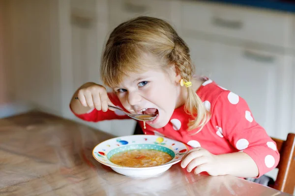 Adorável menina criança comendo sopa de legumes cozidos frescos na cozinha. Criança feliz come comida saudável ao almoço ou jantar. Aprendizagem de bebés. Casa, berçário, escola infantil ou creche — Fotografia de Stock