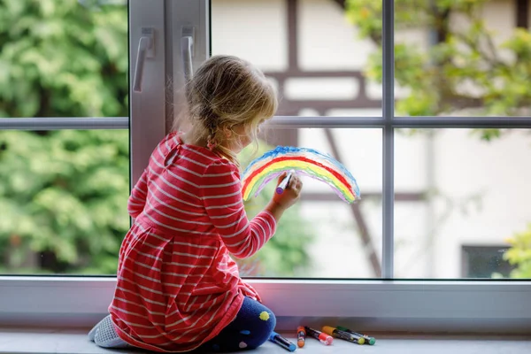 Adoralbe liten småbarn flicka med regnbåge målad med färgglada fönster färg under pandemisk coronavirus karantän. Barnmålning regnbågar och hjärtan runt om i världen med ord Låt alla må bra. — Stockfoto