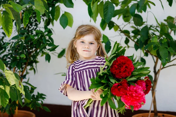 Schattig schattig klein peuter meisje met enorme boeket bloeiende rode en roze pioenroos bloemen. Portret van een lachend kleuter in de binnentuin op warme lente- of zomerdag. Zomertijd. — Stockfoto