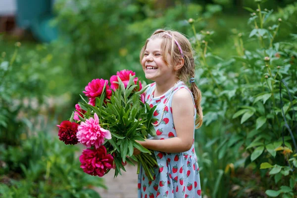 可愛い赤とピンクの牡丹の花の巨大な花束を持つかわいいかわいい幼児の女の子。暖かい春や夏の日に家庭菜園で笑顔の就学前の子供の肖像画。夏. — ストック写真