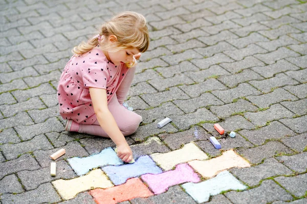 Kleine kleuter meisje schilderen met kleurrijke krijtjes op de grond op de achtertuin. Positief gelukkig peuter kind tekenen en het maken van foto 's. Creatieve buitenactiviteiten in de zomer. — Stockfoto