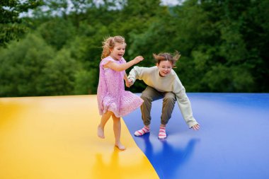 Little preschool girl and school sister jumping on trampoline. Happy funny children, siblings in love having fun with outdoor activity in summer. Trampolin in ukrainian flagg colors. clipart