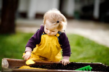 Bahar günü kum ve kürekle oynayan sevimli, tapılası bir kız. Sarı çizme ve çamur yağmurlu su birikintisi pantolon giyen bebek. Baharda sebze eken mutlu kız.