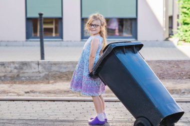 Gözlüklü küçük anaokulu kızı çöp tenekesi alıyor. Çocuk atık ayıklamayı öğreniyor. Çevre kavramı
