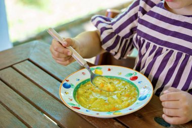 Little preschool girl eating from spoon vegetable potato soup. Healthy food for children. Happy kid at home or nursery