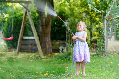 Mutlu küçük kız sıcak ve güneşli bir yaz akşamında bahçe hortumuyla oynuyor. Anaokulu çocuğu evcil bahçedeki ağaçları ve bitkileri sulayarak eğleniyor ve yardım ediyor..