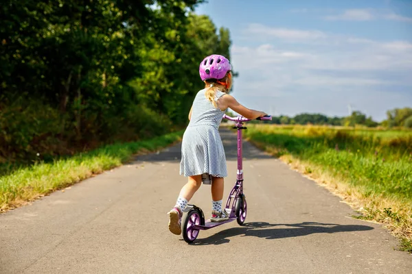 Portrait of active little preschool girl riding scooter on road in park outdoors on summer day. Seasonal child activity sport. Healthy childhood lifestyle.