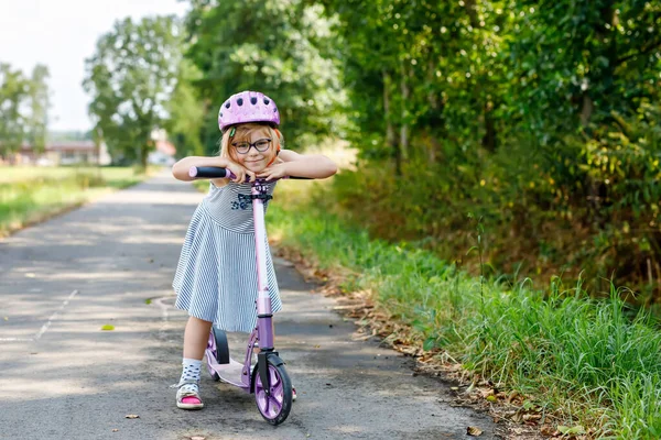 Portrait of active little preschool girl with glasses and helmet riding scooter on road in park outdoors on summer day. Seasonal child activity sport. Healthy childhood lifestyle.
