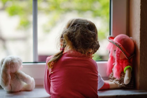 かわいい幼児の女の子は窓のそばに座って雨の日を見ています 人形やおもちゃの孤独感を持つ夢の子供 コロナウイルスパンデミック時の自己分離概念 顔も認識もできない — ストック写真