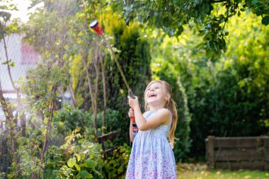 Mutlu küçük kız sıcak ve güneşli bir yaz akşamında bahçe hortumuyla oynuyor. Anaokulu çocuğu evcil bahçedeki ağaçları ve bitkileri sulayarak eğleniyor ve yardım ediyor..