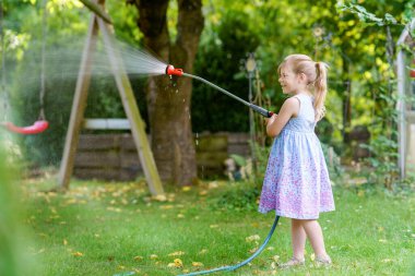 Mutlu küçük kız sıcak ve güneşli bir yaz akşamında bahçe hortumuyla oynuyor. Anaokulu çocuğu evcil bahçedeki ağaçları ve bitkileri sulayarak eğleniyor ve yardım ediyor..