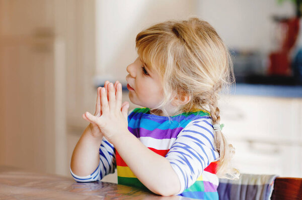 Cute toddler girl praying to God at home. Child using hands for pray and thank for food. Christian tradition