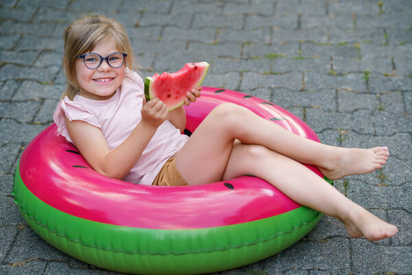 Cute little girl with glasses eating watermelon on inflatable ring in summertime. Happy smiling preschool chil having fun. Healthy summer food and snacks for kids.