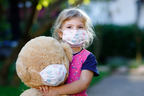 Little toddler girl in medical mask as protection against pandemic coronavirus quarantine disease. Cute child using protective equipment as fight against covid 19 and holding big bear toy with mask