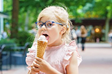 Little preschool girl with glasses eating ice cream in waffle cone on sunny summer day. Happy toddler child eat icecream dessert. Sweet food on hot warm summertime days.