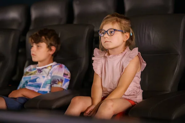 Little preschool girl with glasses and kid boy watching cartoon movie in cinema and eating popcorn. Happy excited children, siblings, brother and sister. Family activity with children