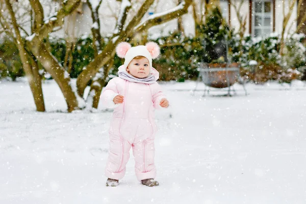 Happy little baby girl making first steps outdoors in winter through snow. Cute toddler learning walking. Child having fun on cold snowy day. Wearing warm baby pink clothes snowsuit and bobbles hat