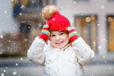 Kış kıyafetleri içinde küçük bir anaokulu kızının komik portresi. Açık havada kırmızı şapkalı ve renkli eldivenli mutlu bir çocuk. Kış günü