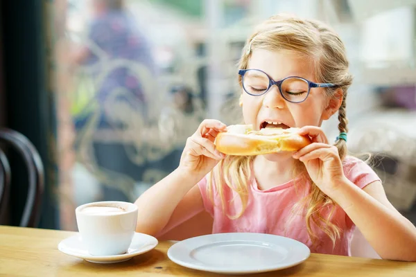 little smiling girl with glasses have a breakfast in a cafe. Preschool child with glasses drinking milk and eating bakery pastry croissant or cake. Happy children, healthy food and meal