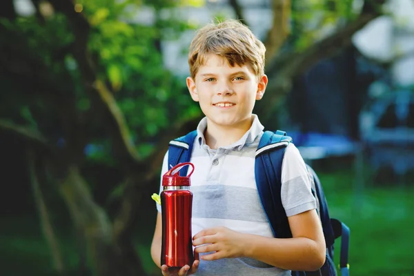 Happy little kid boy drinking from water bottle and backpack or satchel. Schoolkid on way to school. Healthy child outdoors. Back to school