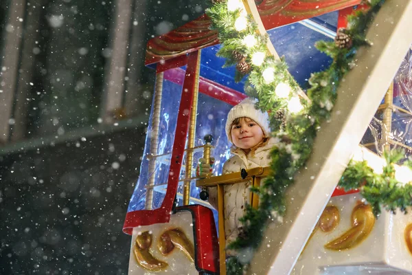 Little preschool girl riding on ferris wheel carousel horse at Christmas funfair or market, outdoors. Happy toddler child having fun on traditional family xmas market in Germany.