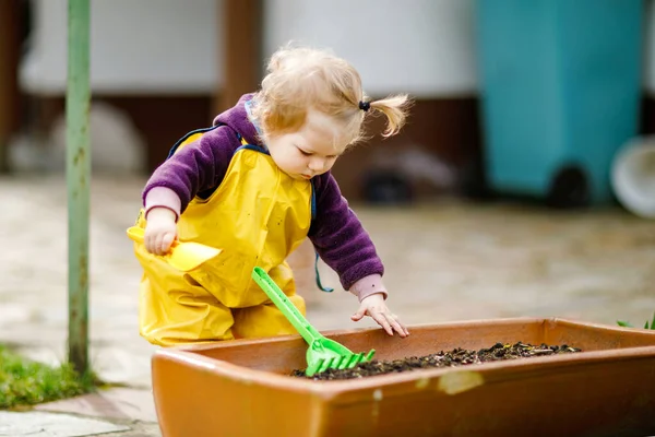 春の日に砂とシャベルで遊んでかわいい愛らしい幼児の女の子 黄色のブーツと泥の雨の水たまりのズボンを着て赤ちゃん 春に野菜を植える幸せな女の子 — ストック写真