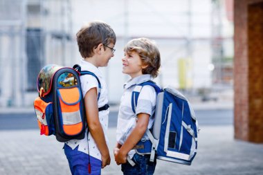 Sırt çantası ya da çantası olan iki küçük çocuk. Okulun yolu üzerindeki okul çocukları. Sağlıklı sevimli çocuklar, erkek kardeşler ve en iyi arkadaşlar sokakta evden ayrılıyor. Okula dönelim. Mutlu kardeşler.