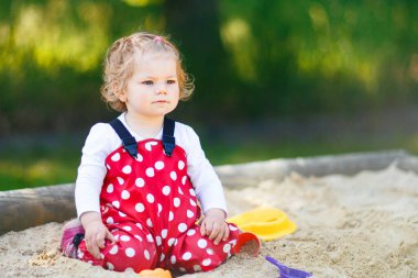 Dışarıda parkta kumlarda oynayan tatlı bir kız çocuğu. Kırmızı sakız pantolonlu güzel bebek güneşli yaz gününde eğleniyor. Renkli kum oyuncakları olan çocuk. Sağlıklı aktif bebek dışarıda oyunlar oynuyor.