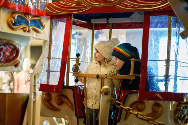 Little preschool girl and school boy riding on ferris wheel carousel horse at Christmas funfair or market, outdoors. Two happy children having fun on traditional family xmas market in Germany.