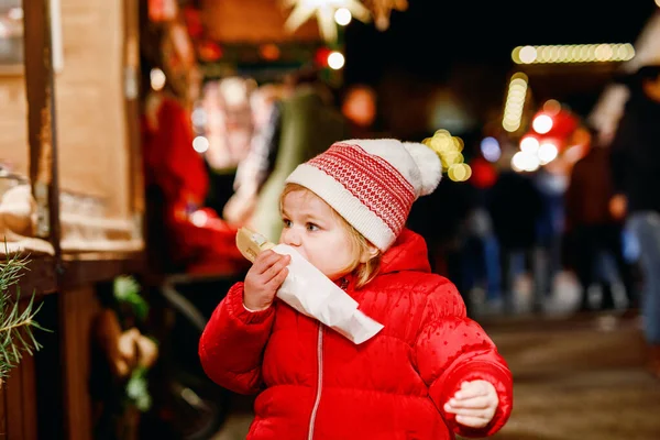 小さな女の赤ちゃん かわいい子がバナナを食べては チョコレート Marshmellows ジンジャーブレッド ナッツと甘いスタンド近くカラフルなスプリンクルで覆われています ドイツのクリスマス マーケットで幸せな幼児 — ストック写真