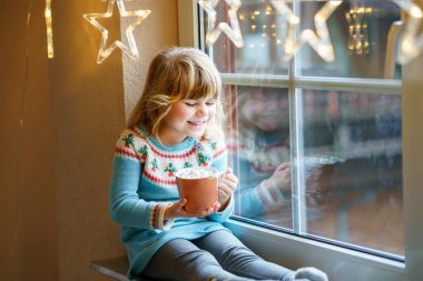 Küçük anaokulu kızı elinde şekerlemeli sıcak çikolata bardağıyla. Mutlu çocuk kışın Noel ışıklarıyla pencerenin yanında tatlı kakao içiyor. Noel 'in samimi aile kutlaması