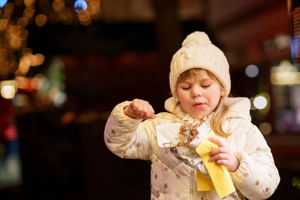 小さな就学前の女の子 かわいい子供が背景に装飾や照明でチョコレートで覆われたチュロスのお菓子を食べています ドイツのクリスマスマーケットで幸せな子供 — ストック写真
